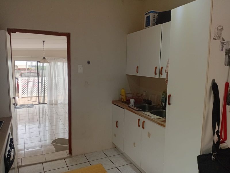 A compact kitchen area featuring white built-in cabinetry, a double bowl sink, and tiled flooring, with an open doorway leading to an adjacent living space.
