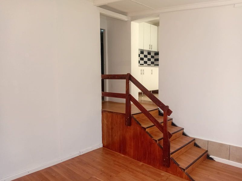 An empty living area featuring a wooden staircase with a handrail, leading towards a kitchen with white cabinetry and a checkered backsplash.