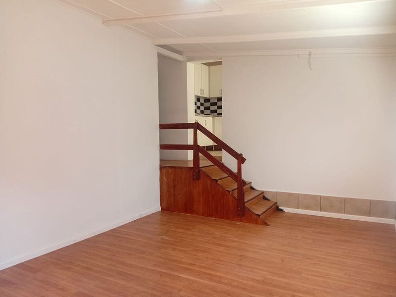 An empty living area featuring laminate flooring and a wooden staircase leading to an adjacent kitchen space.
