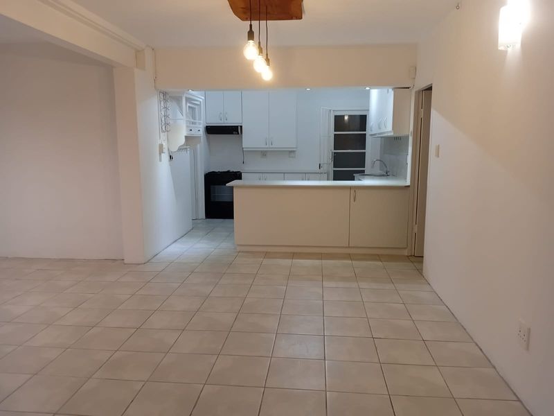 An open-plan living and kitchen area featuring neutral tiled flooring, white cabinetry, a breakfast counter, and built-in kitchen appliances.