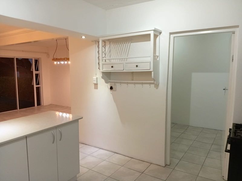 A bright, open-plan kitchen area featuring tiled flooring, a central island, and built-in wall shelving, leading into an adjacent living space.