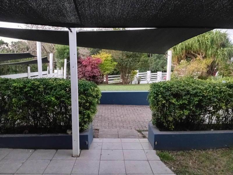 A covered outdoor patio area featuring paved flooring, shade sails, and landscaped garden beds with a white perimeter fence.