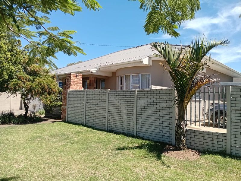 Front exterior view of a single-story house featuring a brick boundary wall, metal security fencing, and a corrugated metal roof.