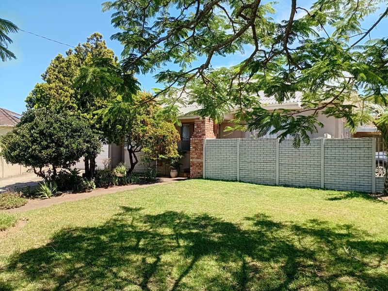 Front view of the property featuring a well-maintained lawn, mature trees, a brick boundary wall, and a paved driveway leading to the entrance.