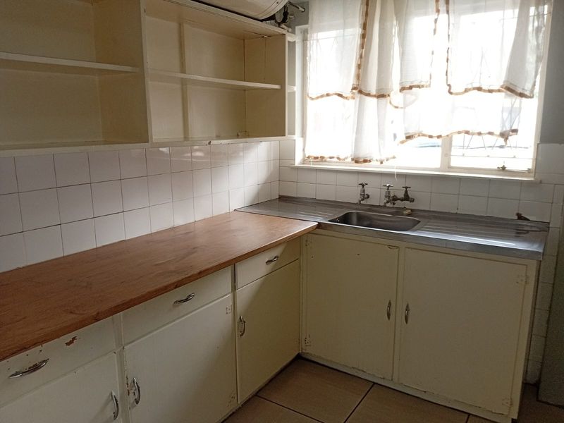 A dated kitchen featuring built-in cabinetry, a laminate countertop, and a stainless steel sink with tiled backsplash and flooring.