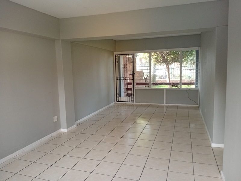 A spacious, empty living area featuring neutral grey walls, tiled flooring, and a large window with security bars and a security gate.