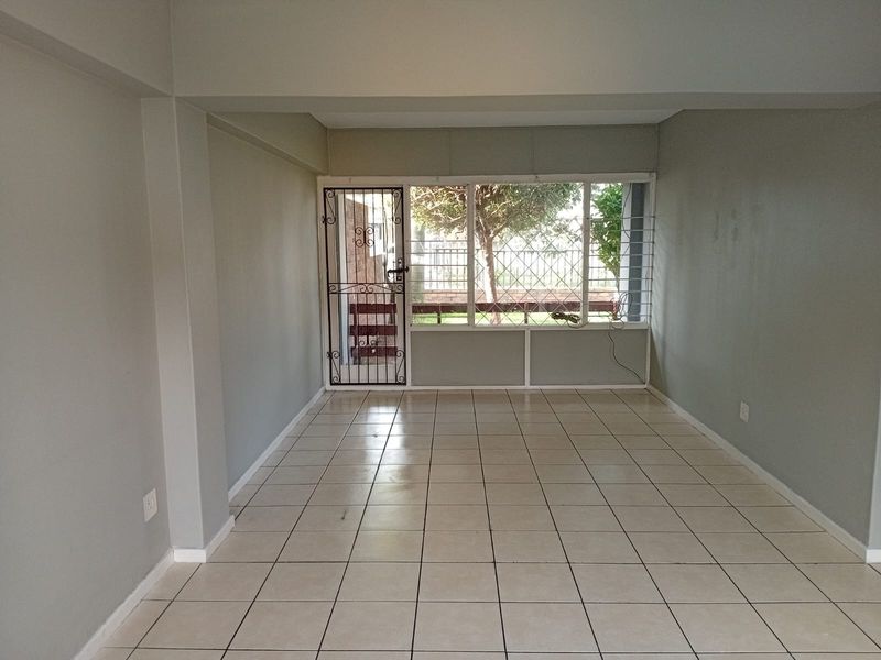 An empty, open-plan living area featuring neutral tiled flooring, large windows with security bars, and a secure metal entrance gate.