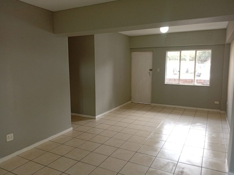 A spacious, empty living area featuring neutral wall paint, tiled flooring, and natural light from a large window.