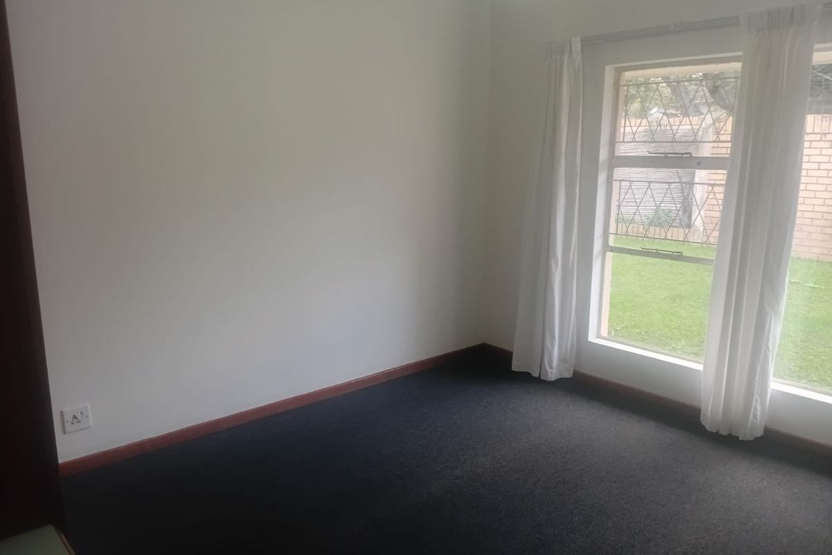 A vacant bedroom featuring dark carpeting, neutral walls, and a window with security bars overlooking the garden area.