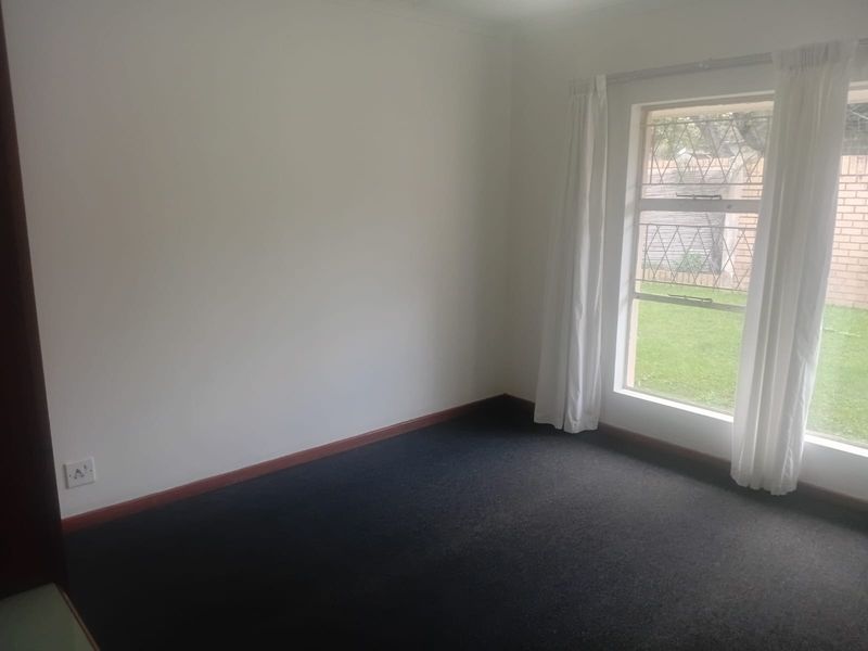 A vacant bedroom featuring dark carpeting, neutral walls, and a window with security bars overlooking the garden area.