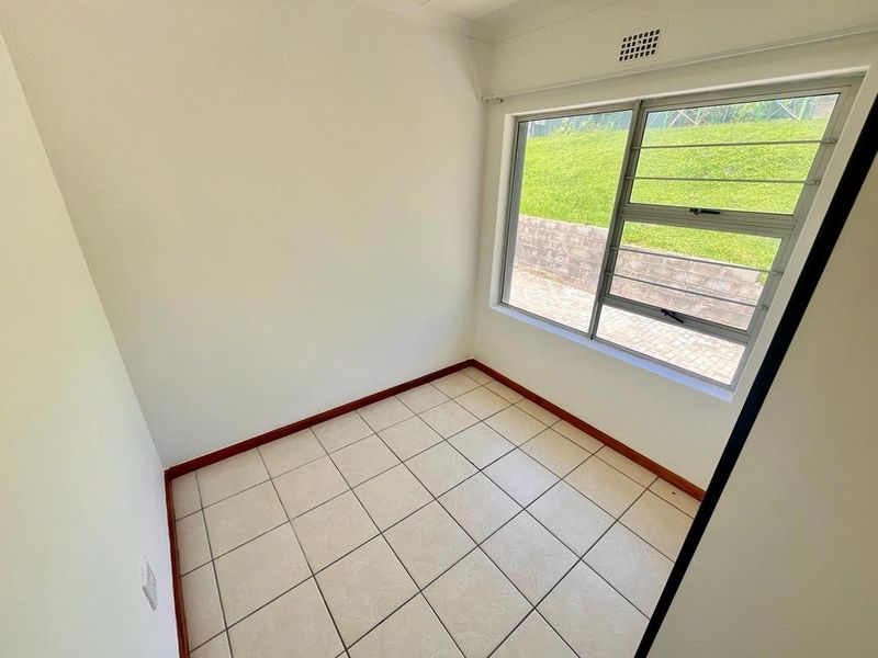A bright, empty bedroom featuring neutral tiled flooring and a large aluminium window with security bars, offering a view of the exterior garden area.