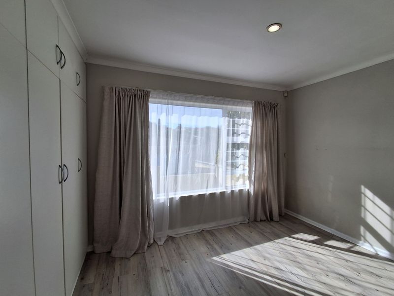 A bright, empty bedroom featuring modern laminate flooring, built-in cupboards, and large windows with neutral curtains.