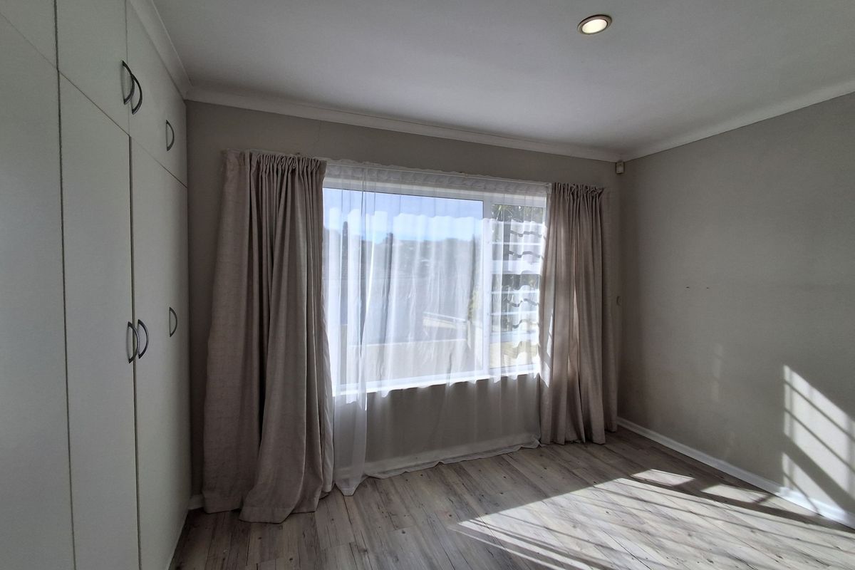 A bright, empty bedroom featuring modern laminate flooring, built-in cupboards, and large windows with neutral curtains.