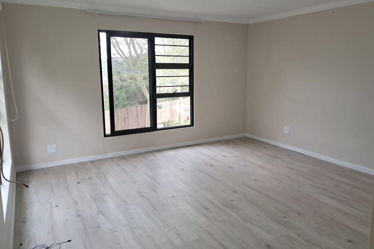 A bright, empty living room featuring modern light-toned laminate flooring, neutral walls, and a large window with security bars.