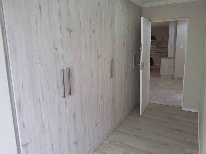 A clean, empty bedroom featuring modern light wood-look built-in cupboards and matching laminate flooring, with a view into the adjacent kitchen area.