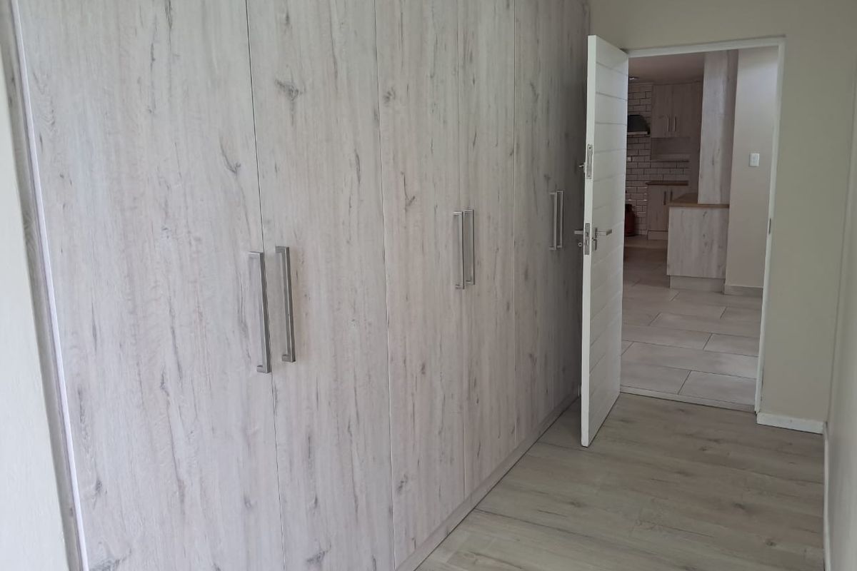 A clean, empty bedroom featuring modern light wood-look built-in cupboards and matching laminate flooring, with a view into the adjacent kitchen area.