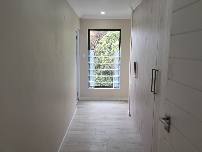 A bright, modern hallway featuring light wood-look laminate flooring, extensive built-in storage cupboards, and a unique glass block window detail.