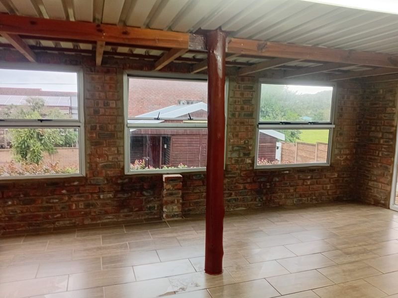 An empty living area featuring exposed brick walls, tiled flooring, and large windows, currently in an unfinished state with exposed ceiling beams.