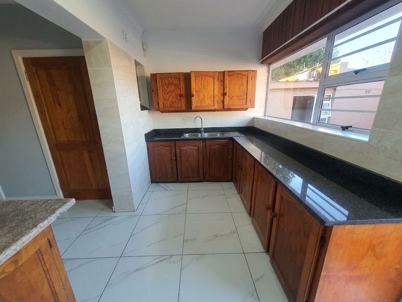 A functional kitchen featuring wooden cabinetry, granite countertops, a stainless steel sink, and modern white tiled flooring with a large window.