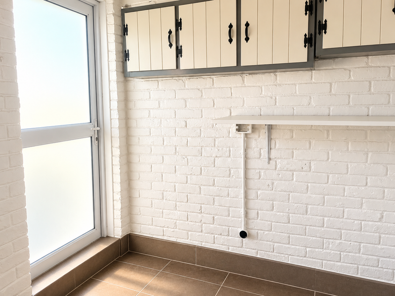 A clean and bright laundry area featuring white painted brick walls, built-in overhead cabinetry, fixed shelving, and durable tiled flooring with direct exterior access.