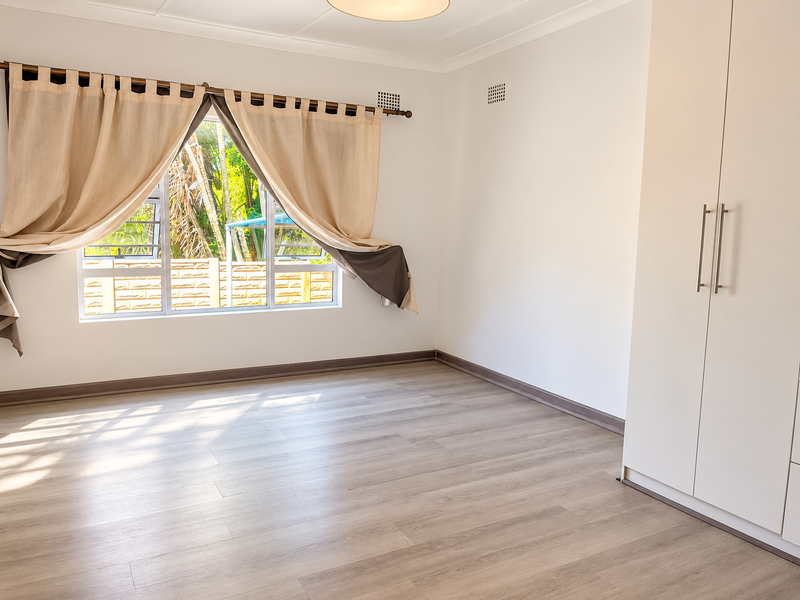 A bright, empty bedroom featuring modern laminate flooring, a built-in wardrobe, and large windows with natural light.