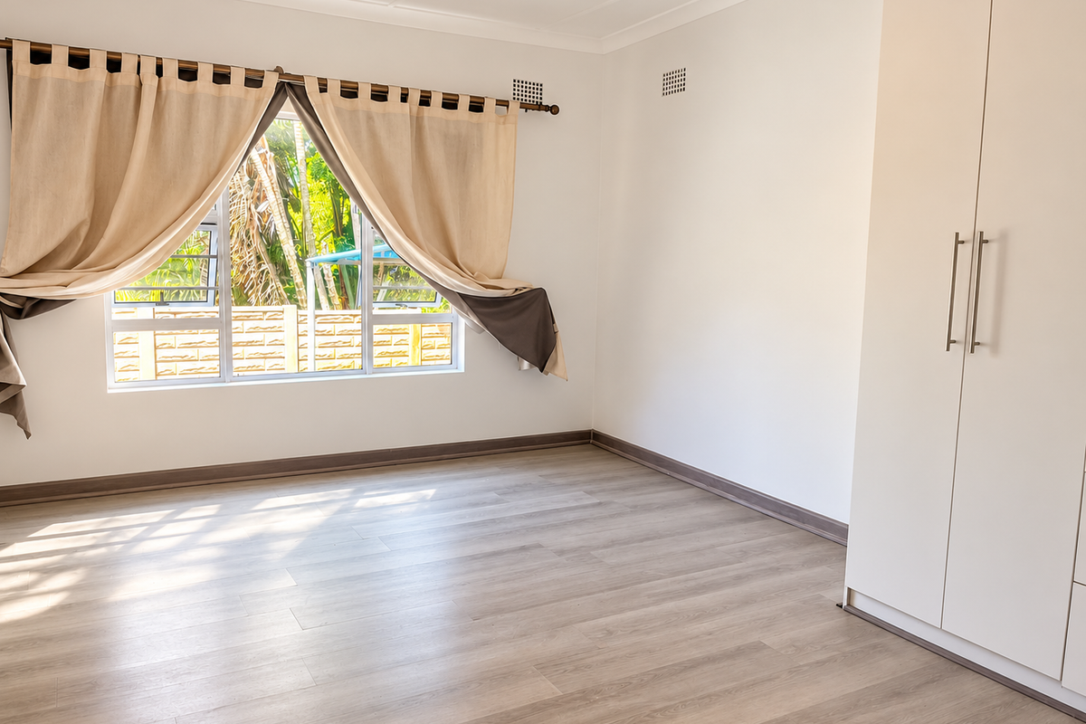 A bright, empty bedroom featuring modern laminate flooring, a built-in wardrobe, and large windows with natural light.