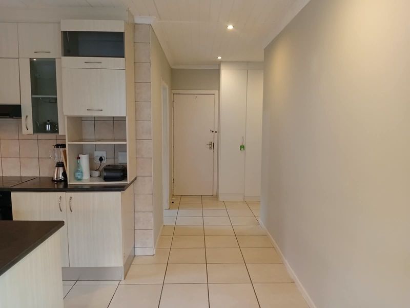 A functional kitchen area featuring light-toned built-in cabinetry, tiled flooring, and a tiled backsplash, leading into a hallway with additional storage cupboards.