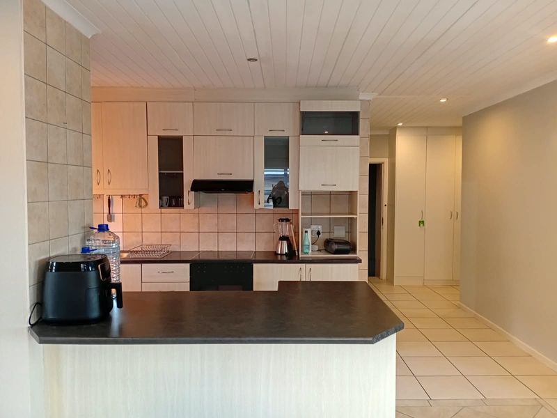 Functional kitchen featuring light-toned cabinetry, dark laminate countertops, integrated appliances, and a breakfast bar, leading into a tiled hallway with built-in storage.