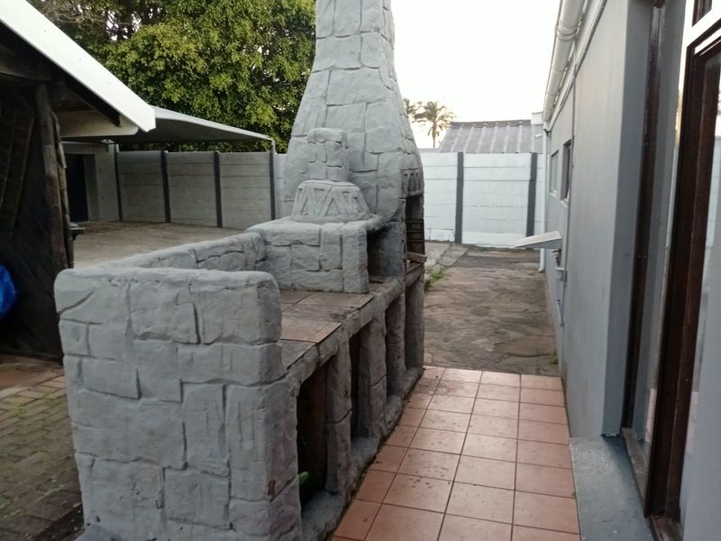 Rear exterior area featuring a large built-in stone braai, tiled patio space, and paved walkway enclosed by precast boundary walls.