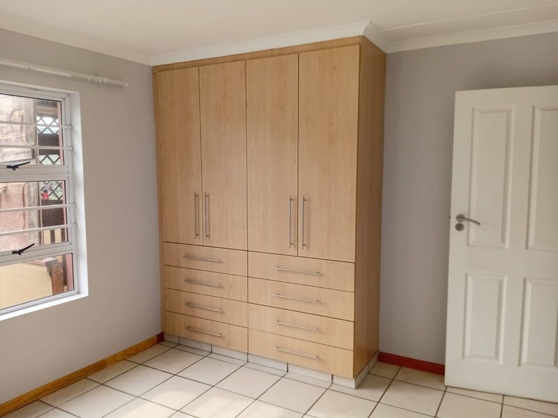 A clean, empty bedroom featuring light wood-finish built-in cupboards, neutral wall paint, tiled flooring, and a modern aluminium window frame.