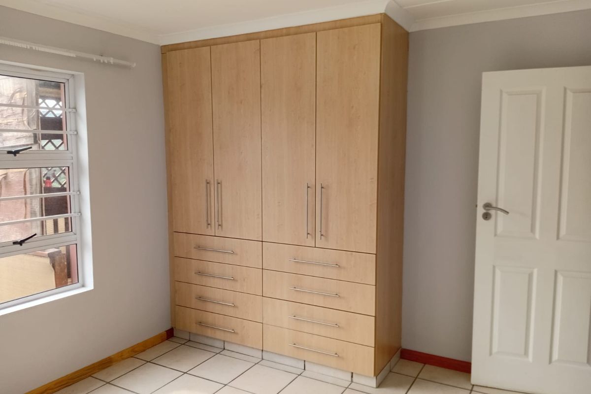 A clean, empty bedroom featuring light wood-finish built-in cupboards, neutral wall paint, tiled flooring, and a modern aluminium window frame.