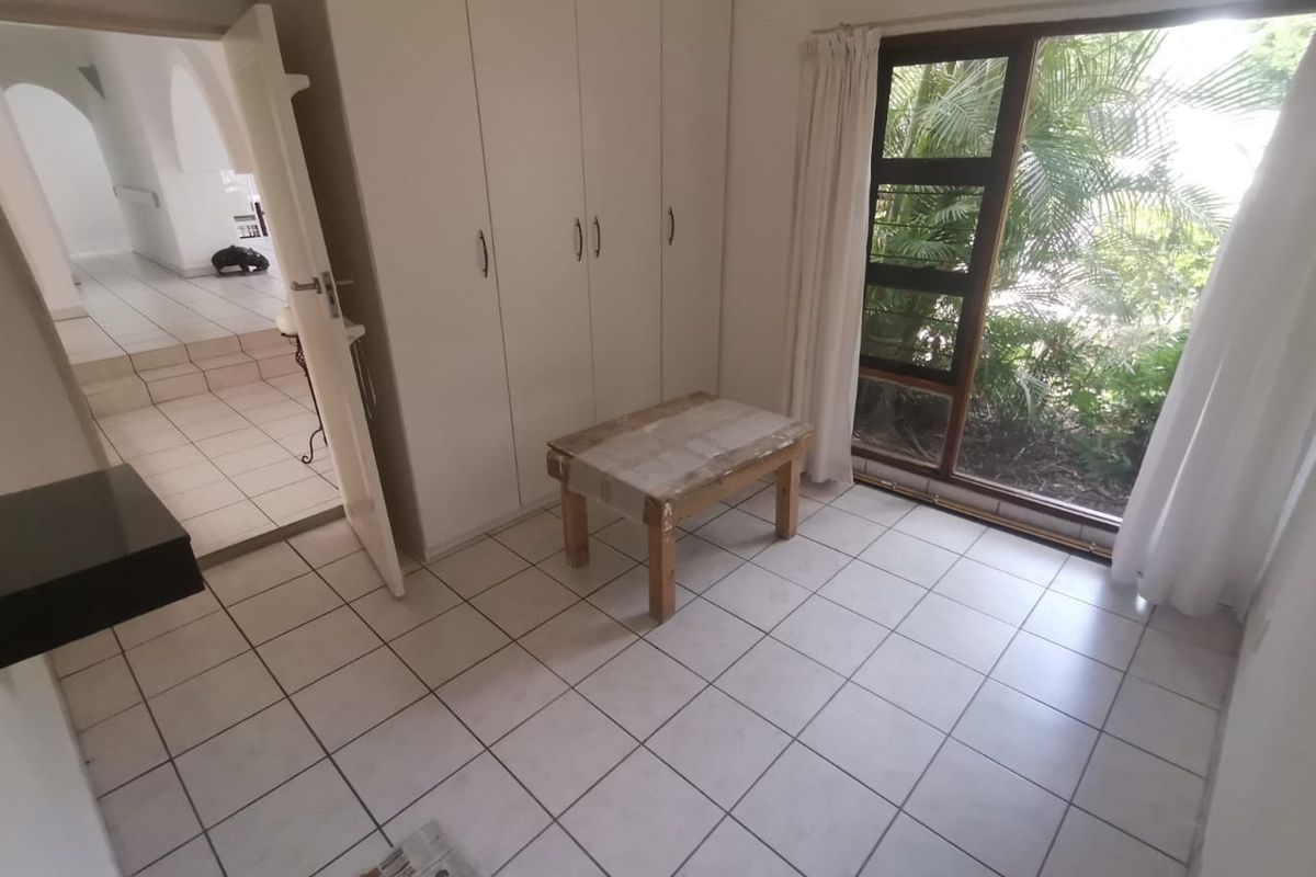 A bright, empty bedroom featuring tiled flooring, built-in cupboards, and a large window overlooking the garden.