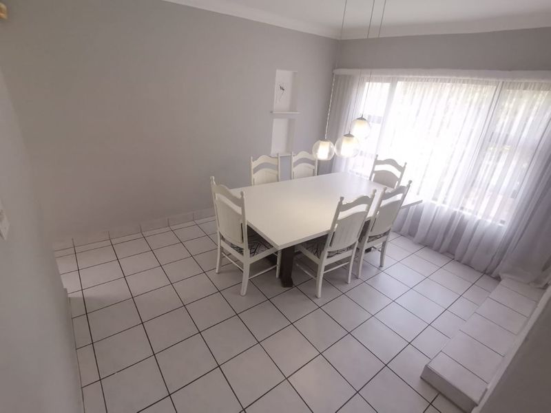 A bright dining room featuring tiled flooring, a recessed wall niche, and modern pendant lighting, illuminated by a large window with sheer curtains.