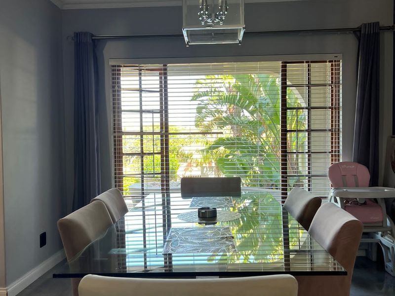 A well-lit dining room featuring tiled flooring, a modern pendant light fixture, and large windows with venetian blinds overlooking the garden.