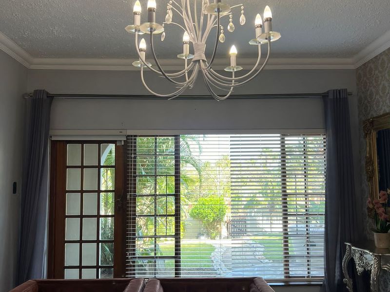 A well-lit living room featuring a decorative chandelier, tiled flooring, and a large window with venetian blinds providing a view of the garden.