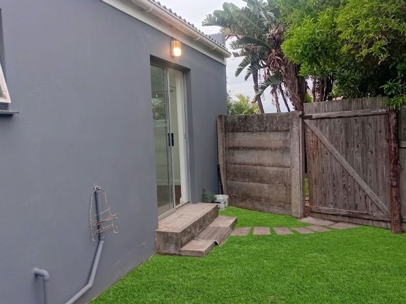 Rear exterior view of the property featuring a sliding glass door, concrete steps, a small lawn, and a wooden gate with precast boundary walling.