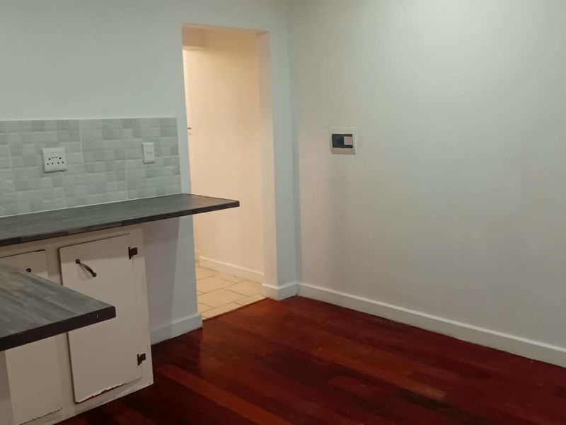 A functional kitchen area featuring hardwood flooring, laminate countertops, and a tiled backsplash, leading into an adjacent utility space.