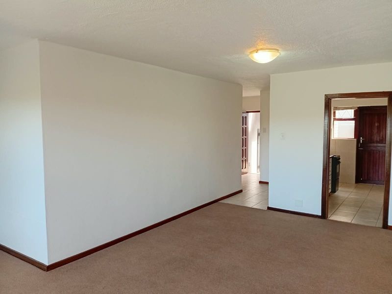 An empty living room area featuring neutral carpeting, white walls, and access to an adjacent tiled hallway and kitchen area.