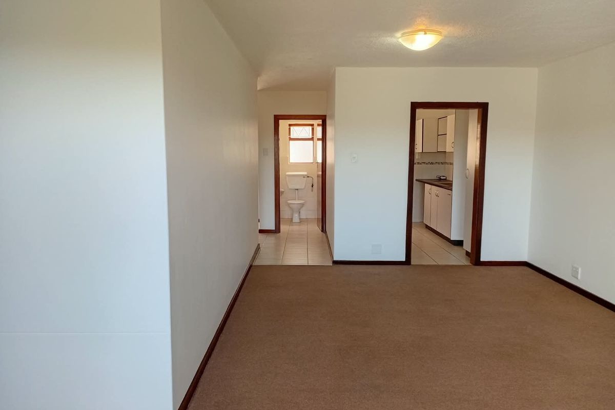 An empty living area featuring neutral carpeting, leading into a kitchen and bathroom with tiled flooring and timber door frames.