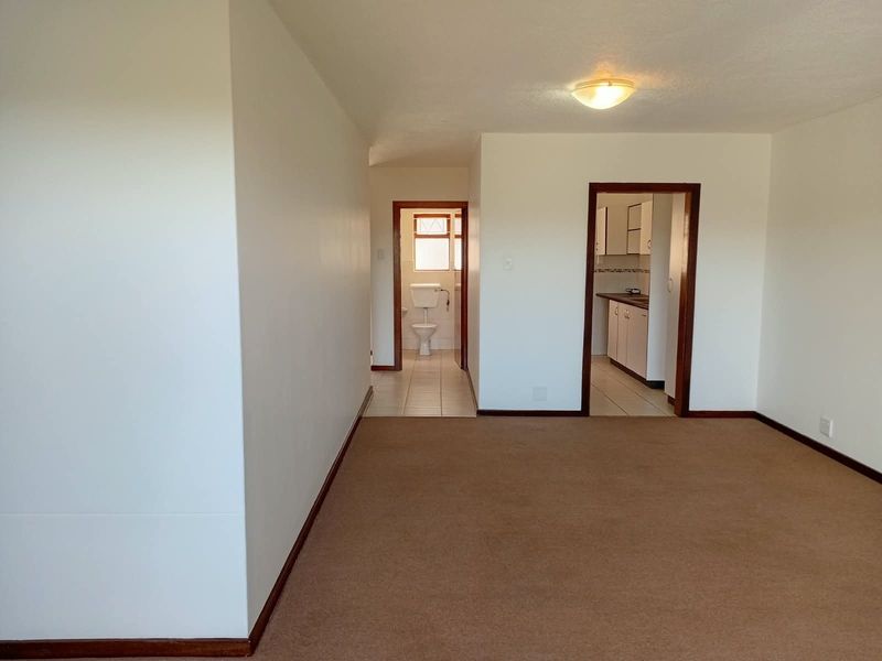 An empty living area featuring neutral carpeting, leading into a kitchen and bathroom with tiled flooring and timber door frames.