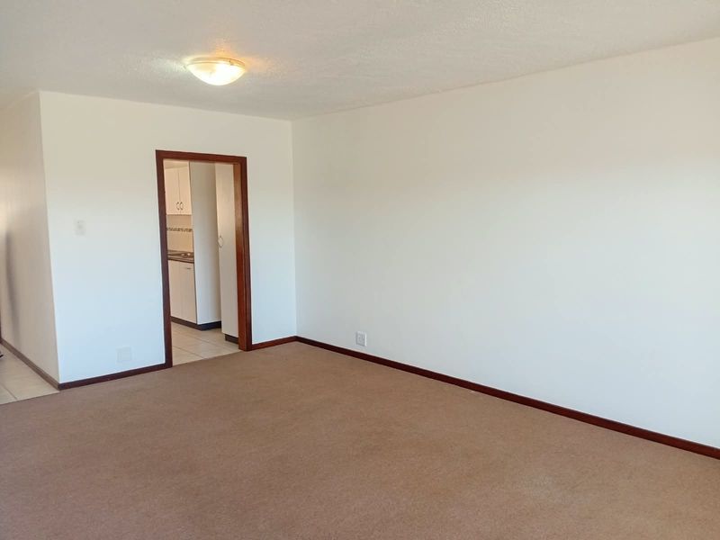 A spacious, empty living room featuring neutral carpet flooring and a doorway leading to the kitchen area.