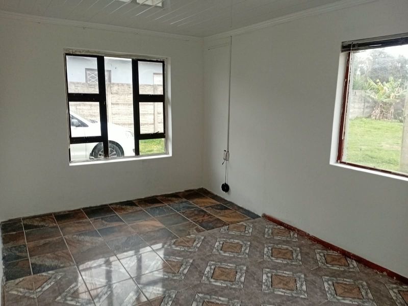 An empty living room featuring mismatched tiled flooring, aluminium window frames, and a tongue and groove ceiling, requiring cosmetic updates.