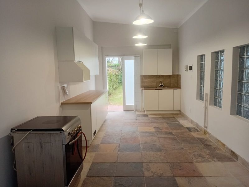 A bright, empty kitchen featuring stone tile flooring, white cabinetry, glass block windows, and direct access to the rear garden.