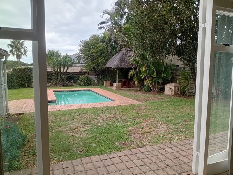 A private backyard featuring a swimming pool, a paved patio area, and a traditional thatched lapa set within a landscaped garden.