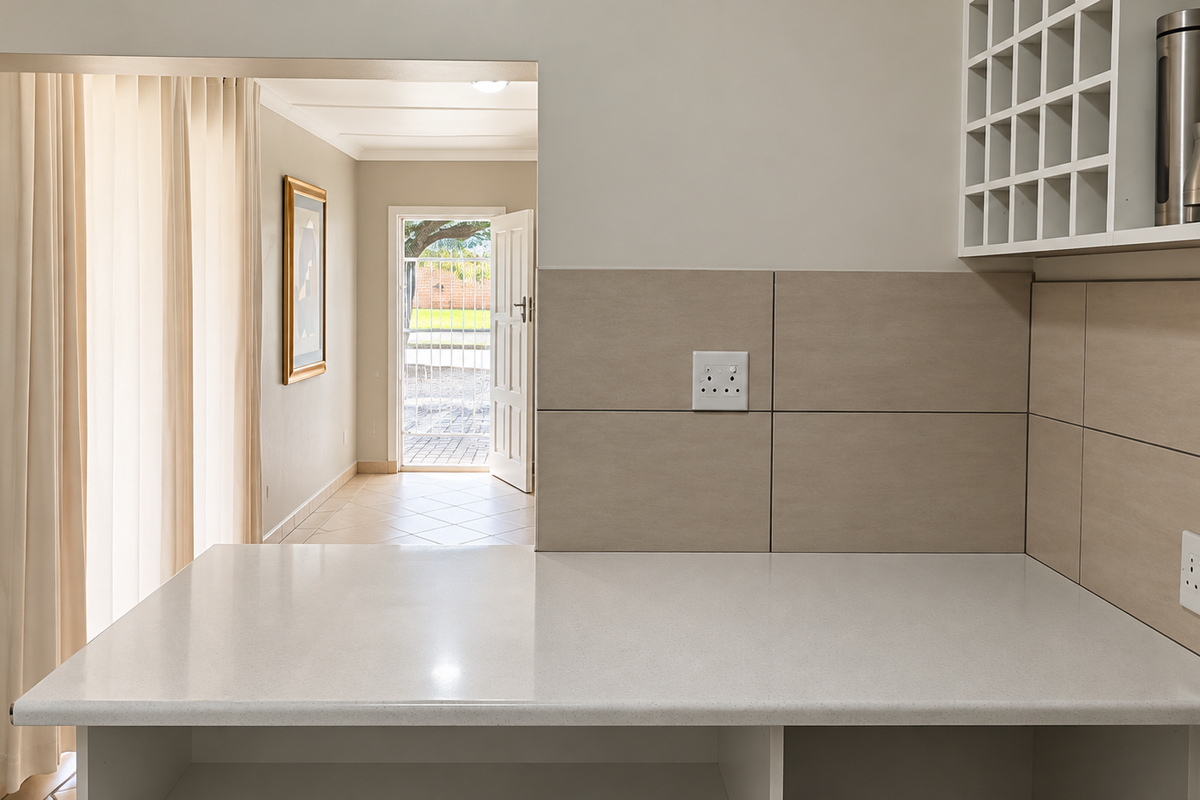 A clean kitchen area featuring a stone countertop, tiled splashback, and built-in wine storage, with a view through to the entrance hall.