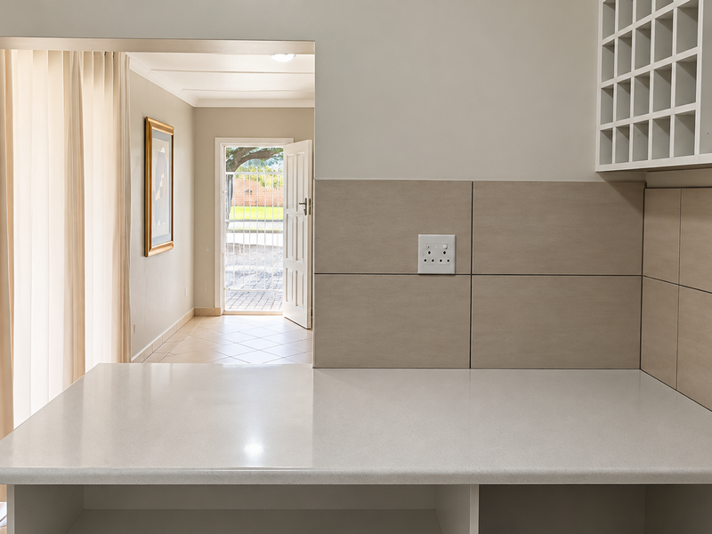 A clean kitchen area featuring a stone countertop, tiled splashback, and built-in wine storage, with a view through to the entrance hall.