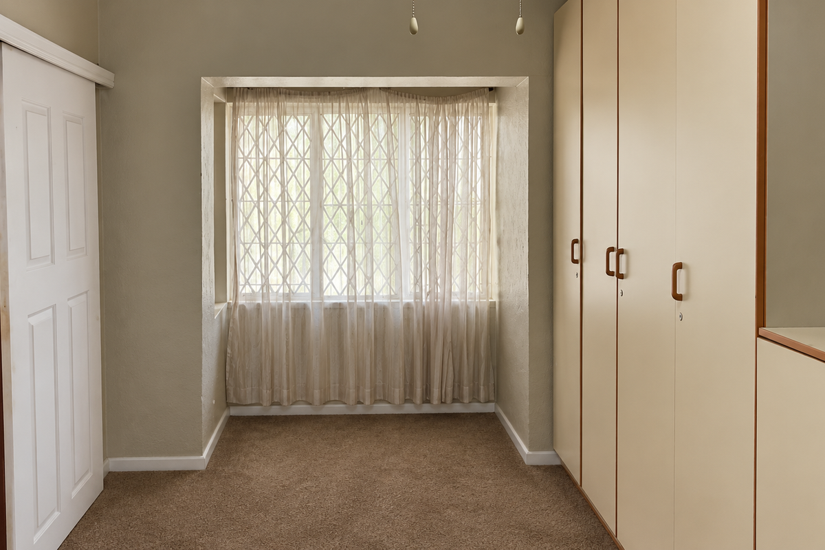 A clean, empty bedroom featuring neutral walls, carpeted flooring, and extensive built-in cupboard storage with a window fitted with a security gate.