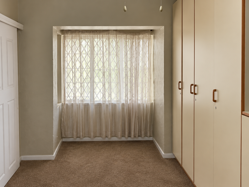 A clean, empty bedroom featuring neutral walls, carpeted flooring, and extensive built-in cupboard storage with a window fitted with a security gate.