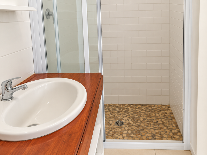 A clean, modern bathroom featuring a wooden vanity with a white basin and a walk-in shower with subway tiles and a pebble floor.