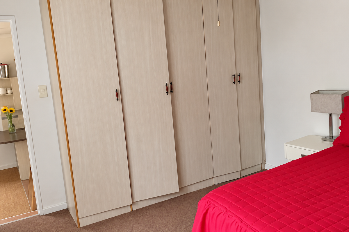 A bedroom featuring light wood-finish built-in wardrobes and carpeted flooring, with a view into an adjacent room.